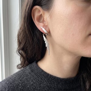 May include: A silver-toned ear cuff shaped like a pair of wings. The upper wing curves along the top of the ear cartilage, while the lower wing extends downward along the earlobe, featuring detailed feather-like engravings on the metal surface.