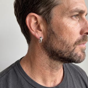 Peut inclure: Une petite boucle d'oreille créole en argent représentant un cobra détaillé. La boucle d'oreille est portée au lobe et présente une finition métallique polie avec des écailles texturées le long du corps du serpent, formant un anneau circulaire.