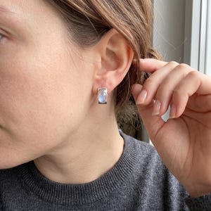 May include: A silver stud earring featuring a smooth, oval-shaped moonstone cabochon set within a rectangular metal frame. The stone displays a soft, iridescent blue glow against the polished silver setting, worn on the earlobe.