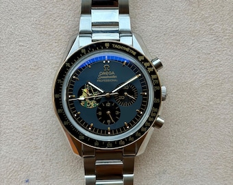 May include: A silver-tone stainless steel Omega Speedmaster Professional chronograph watch with a black dial, tachymeter bezel, and a metallic bracelet. The dial features three sub-dials, gold-tone hands, and a small colorful graphic illustration.