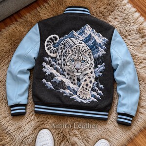 Puede incluir: Una chaqueta tipo varsity con cuerpo gris oscuro y mangas azul claro. La espalda presenta un gráfico detallado de un leopardo de las nieves en un paisaje montañoso. El cuello, los puños y el dobladillo tienen rayas negras y azul claro. Texto: Amiro Leather.
