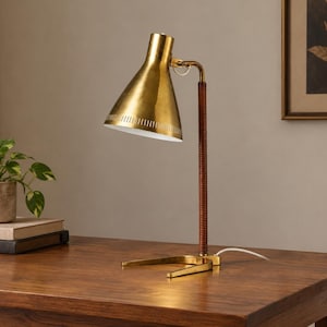 Puede incluir: Una lámpara de escritorio con pantalla cónica de latón, interior blanco y borde perforado. El vástago vertical está revestido en cuero marrón y se conecta a una base de latón con dos patas curvas. Un cable de alimentación blanco sale de la base.