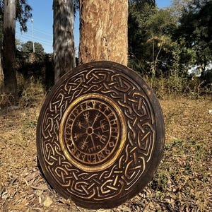 Könnte beinhalten: Eine kreisförmige Wandplatte aus Holz mit dunkelbraunem Finish und aufwendigen, tief geschnitzten keltischen Knotenmustern, die ein zentrales Medaillon umgeben. Die Mitte zeigt eine helle Holzeinlage mit einem schwarzen Vegvisir-Symbol und Runen.