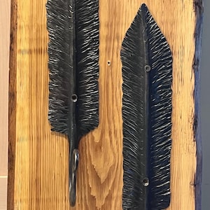 May include: Two black metal wall hooks shaped like textured feathers, each featuring a curved hook at the bottom and two visible screw holes along the central spine, mounted vertically on a light brown wooden board with a natural grain pattern.