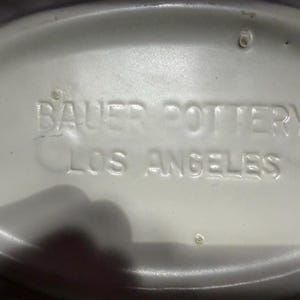 May include: An oval-shaped white ceramic base featuring the embossed text BAUER POTTERY LOS ANGELES centered on the surface. The piece has a smooth, glossy finish with a raised rim around the perimeter and small circular indentations visible.