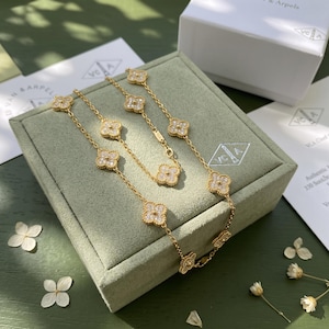 May include: A gold chain necklace featuring multiple four-leaf clover motifs, each encrusted with small clear stones. The necklace is displayed on a sage green velvet jewelry box, with a white branded box and paper cards visible in the background.
