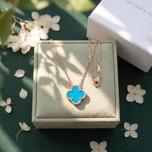May include: A rose gold necklace featuring a four-leaf clover pendant with a bright turquoise inlay. The delicate chain includes a lobster clasp closure. The jewelry is displayed on a square, light green velvet box next to a white gift box.