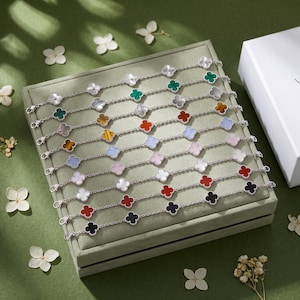 May include: A collection of silver-toned chain bracelets, each featuring multiple four-leaf clover motifs inlaid with various coloured stones including black, red, white, blue, pink, green, and tiger's eye, arranged in parallel rows on a square tray.
