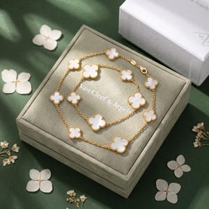 May include: A gold chain necklace and bracelet set featuring multiple white clover-shaped motifs with mother-of-pearl inlays. The jewellery is displayed on a grey velvet box with the text Van Cleef & Arpels printed on the lid in white lettering.