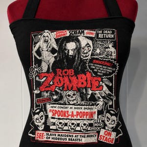 May include: A black halter top featuring a graphic print of Rob Zombie with skulls, text reading "SHOCK SHOW", "SCREAM", "THE DEAD RETURN", "ROB ZOMBIE", "SPOOKS-A-POPPIN", "SEE SLAVE MAIDENS AT THE MERCY OF HIDEOUS BEASTS", and other horror-themed imagery.