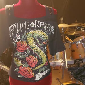 May include: Black off-the-shoulder top with the band name "Falling in Reverse" in white, featuring a green and yellow snake design with red roses. The top is displayed on a red mannequin, with a drum set in the background.