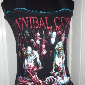 Può includere: Top nero con bretelle e bordino blu con una stampa grafica di una band chiamata "Cannibal Corpse". La grafica presenta una scena sanguinosa con scheletri e un logo della band.