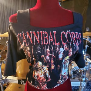 Puede incluir: Camiseta negra con hombros descubiertos con el nombre de la banda "CANNIBAL CORPSE" en letras rojas. La camiseta presenta un gráfico de figuras grotescas. Las mangas son cortas y la camiseta se exhibe en un maniquí rojo.