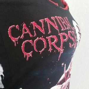 Puede incluir: Una camiseta de tirantes negra con ribete de encaje rojo a lo largo del escote y los tirantes. El frente muestra el texto "CANNIBAL CORPSE" en una fuente roja estilizada sobre una ilustración gráfica de una escena de cementerio con figuras.