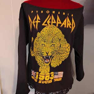Könnte beinhalten: Schwarzes Off-Shoulder-Langarmshirt mit gelbem Leoparden-Grafik und dem Text "Def Leppard Pyromania 1983 United States Tour". Der untere Saum ist mit schwarzer Spitze verziert.