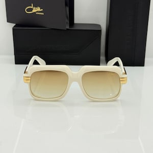 May include: Off-white square-framed sunglasses with gradient brown lenses and gold-tone metal accents at the hinges. The sunglasses are displayed in front of a black branded case and a black card featuring the gold text Cazal Legends.
