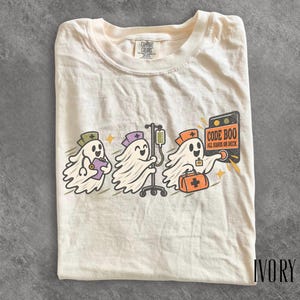 May include: An ivory t-shirt featuring a graphic of three cartoon ghosts dressed as medical staff. One ghost holds a clipboard, one pushes an IV pole, and one carries a medical bag and presses a button labeled CODE BOO ALL HANDS ON DECK.