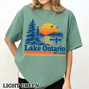 May include: A light green short-sleeve t-shirt featuring a graphic of a lake scene with dark blue pine trees, a yellow and orange sunset, and a person in a canoe. The text reads Lake Ontario EST 1656 in dark blue with orange and yellow stripes.