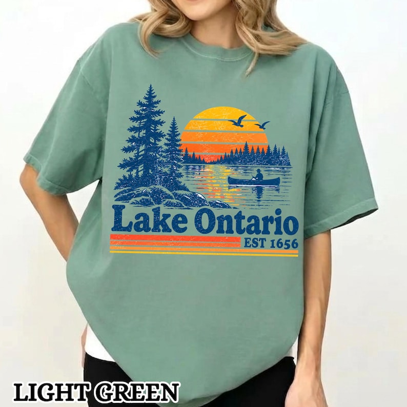 May include: A light green short-sleeve t-shirt featuring a graphic of a lake scene with dark blue pine trees, a yellow and orange sunset, and a person in a canoe. The text reads Lake Ontario EST 1656 in dark blue with orange and yellow stripes.