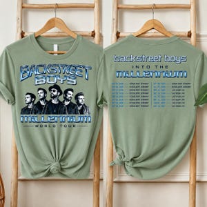 May include: A sage green t-shirt featuring a graphic of the Backstreet Boys with the text "BACKSTREET BOYS INTO THE MILLENNIUM WORLD TOUR" on the front and a list of tour dates and locations printed on the back in blue and white lettering.
