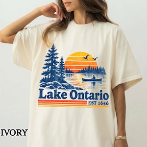 May include: An ivory short-sleeve t-shirt featuring a graphic of Lake Ontario. The design includes a blue pine forest, a sunset with orange and yellow stripes, and a person in a canoe on the water. The text reads Lake Ontario EST 1656.