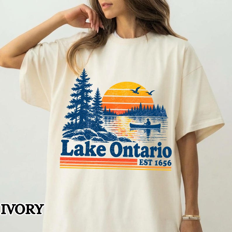 May include: An ivory short-sleeve t-shirt featuring a graphic of Lake Ontario. The design includes a blue pine forest, a sunset with orange and yellow stripes, and a person in a canoe on the water. The text reads Lake Ontario EST 1656.
