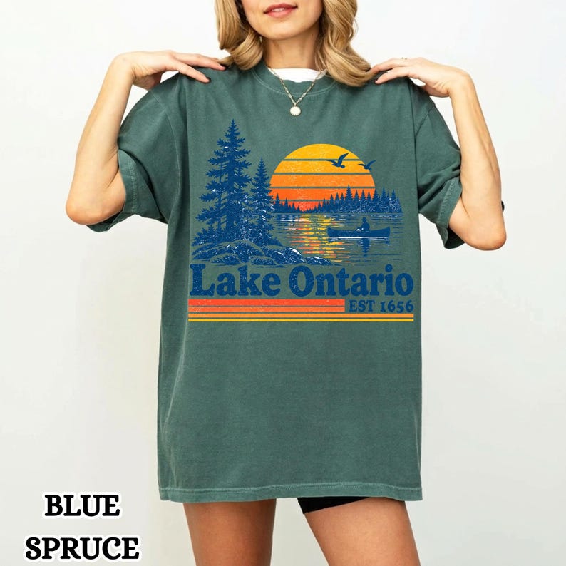 May include: A blue spruce colored short-sleeve t-shirt featuring a graphic print of a lake scene with pine trees, a sunset, and a person in a canoe. The text "Lake Ontario" and "EST 1656" is printed below the graphic in a bold blue font.