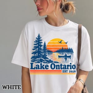 May include: A white t-shirt featuring a graphic print of a sunset over a lake with pine trees, a person in a canoe, and two birds. The text reads Lake Ontario EST 1656 in blue and orange, with horizontal stripes below the text.