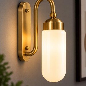 Könnte beinhalten: Eine Wandleuchte mit gebürstetem Messing-Finish, einer vertikalen rechteckigen Rückplatte mit zwei abgerundeten Knöpfen und einem geschwungenen Arm, der einen zylindrischen, mattweißen Glasschirm trägt, welcher ein sanftes Licht abgibt.
