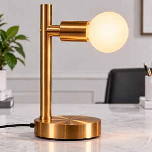 Könnte beinhalten: Eine Tischleuchte mit gebürsteter Goldmetalloberfläche, bestehend aus einem zylindrischen vertikalen Pfosten, einem runden Sockel mit radialem Muster und einem horizontalen Arm, der eine einzelne kugelförmige weiße Glühbirne hält.