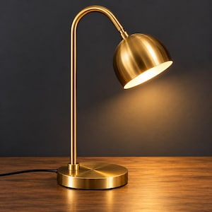 Könnte beinhalten: Eine Schreibtischlampe aus goldfarbenem Metall mit einem schlanken, geschwungenen Hals und einem abgerundeten, kuppelförmigen Lampenschirm. Die Lampe hat einen runden Sockel und strahlt ein warmes, helles Licht von der Unterseite des Schirms aus.