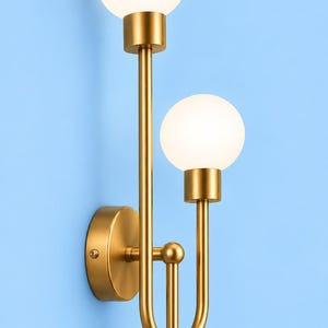 Könnte beinhalten: Eine Wandleuchte mit einem Rahmen aus gebürstetem Goldmetall und zwei vertikalen Armen unterschiedlicher Länge. Jeder Arm ist mit einer kugelförmigen, mattweißen Glaskugel als Leuchtmittel versehen. Die runde Basisplatte dient zur Wandmontage.