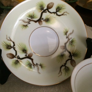 May include: A white porcelain saucer with a gold rim and a delicate design of pine branches and cones in shades of green and brown.
