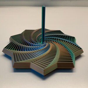 Può includere: Una trottola multicolore a forma di stella con una sfumatura di tonalità verde acqua, blu e marrone chiaro. L'oggetto presenta un manico verticale centrale e un complesso motivo geometrico a strati che si irradia dal centro verso l'esterno.