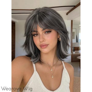 May include: A shoulder-length wig featuring layered, dark gray hair with lighter silver-gray highlights, styled with wispy bangs that frame the forehead. The hair has a textured, voluminous appearance with soft waves throughout the cut.