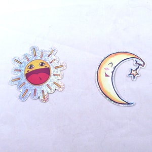 May include: Two colorful stickers, one of a smiling sun with a yellow face and a red mouth, and the other of a smiling crescent moon with a yellow face and a star.