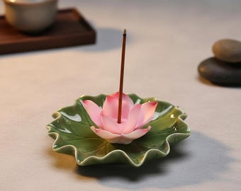 May include: A ceramic incense holder shaped like a green lotus leaf with a pink lotus flower in the centre. A thin, brown incense stick stands upright in the middle of the flower petals. The holder has a glossy finish and wavy, organic edges.
