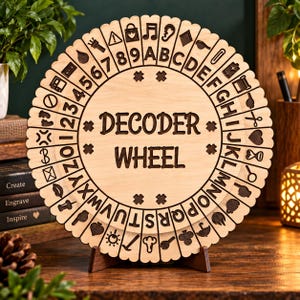May include: A circular wooden decoder wheel featuring engraved text that reads DECODER WHEEL in the center. The outer edge displays a sequence of numbers, letters of the alphabet, and various icons including symbols for music, weather, and tools.