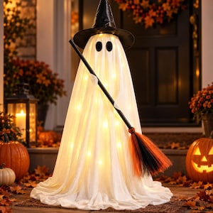 Op de afbeelding: Een witte spookdecoratie met een zwarte puntmuts, verlicht door warm binnenlicht. De spookfiguur houdt een bezem vast met zwarte en oranje borstelharen en heeft twee donkere, ovale ogen op de gedrapeerde, stofachtige buitenkant.