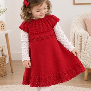 May include: A red knitted sleeveless dress with a ruffled yoke and a textured lace-patterned hem. The dress is worn over a long-sleeved white shirt featuring a subtle light pink polka dot pattern. A small red bow clip is attached to the hair.