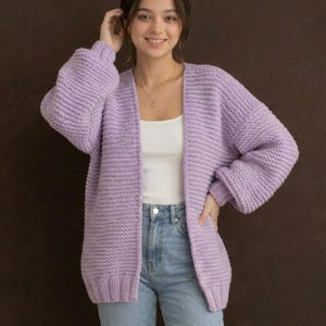 May include: A chunky knit lavender cardigan featuring a relaxed open-front design, long sleeves with ribbed cuffs, and a textured garter stitch pattern. The garment is worn over a simple white ribbed tank top and light blue denim jeans.