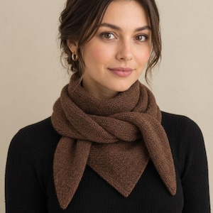 May include: A solid brown knitted triangular scarf worn around the neck, featuring a textured garter stitch pattern with pointed ends, layered over a plain black long-sleeved top. The scarf is wrapped to create a layered, folded appearance.