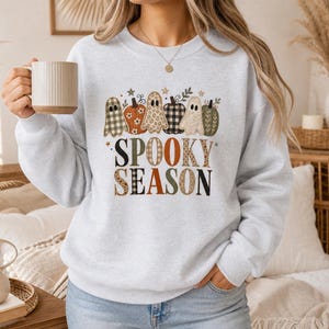 May include: A light gray crewneck sweatshirt featuring a graphic of ghosts and pumpkins with various patterns. The text SPOOKY SEASON is printed below the ghosts and pumpkins in a mix of black, orange, and patterned lettering on the front.