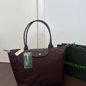Op de afbeelding: Een donkerbruine tote bag met twee dunne hengsels, een klepsluiting met drukknoop en een ritssluiting. De tas is voorzien van een klein in reliëf aangebracht logo aan de voorzijde, een crèmekleurig merklabel en een groen rechthoekig label.