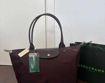 May include: A dark brown tote bag with two thin handles, a snap-button flap closure, and a zipper top. The bag features a small embossed logo on the front, a cream-colored brand tag, and a green rectangular tag attached to the handle.