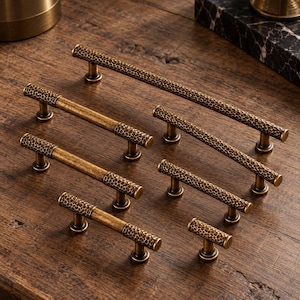 May include: A collection of seven brass cabinet pulls and one knob, each featuring a textured, hammered honeycomb pattern on the ends and a smooth, aged metallic finish. The hardware varies in length, ranging from a single knob to long bars.