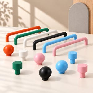 May include: A collection of cabinet hardware including six long, arched bar pulls and eight round knobs in various solid colors: orange, green, white, black, blue, and pink. The hardware pieces are arranged on a smooth, light-colored surface.