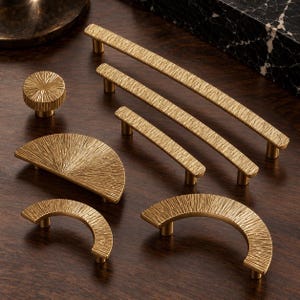 May include: A collection of gold-toned metal cabinet hardware featuring a textured, linear pattern. The set includes one circular knob, two straight bar pulls of varying lengths, one semi-circular pull, and two arched, curved handles.