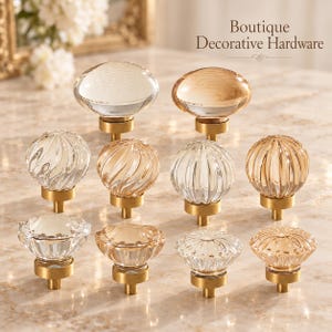 May include: A collection of ten cabinet knobs featuring clear and amber-tinted glass tops with various textures, including smooth, ribbed, and fluted designs, all mounted on brushed brass bases. The text Boutique Decorative Hardware is visible.