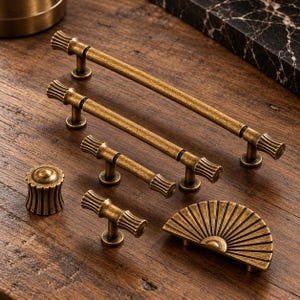 May include: A collection of antique brass cabinet hardware including three varying lengths of cylindrical bar pulls, one round fluted knob, one small T-bar handle, and one semi-circular fan-shaped pull, all featuring a textured finish.